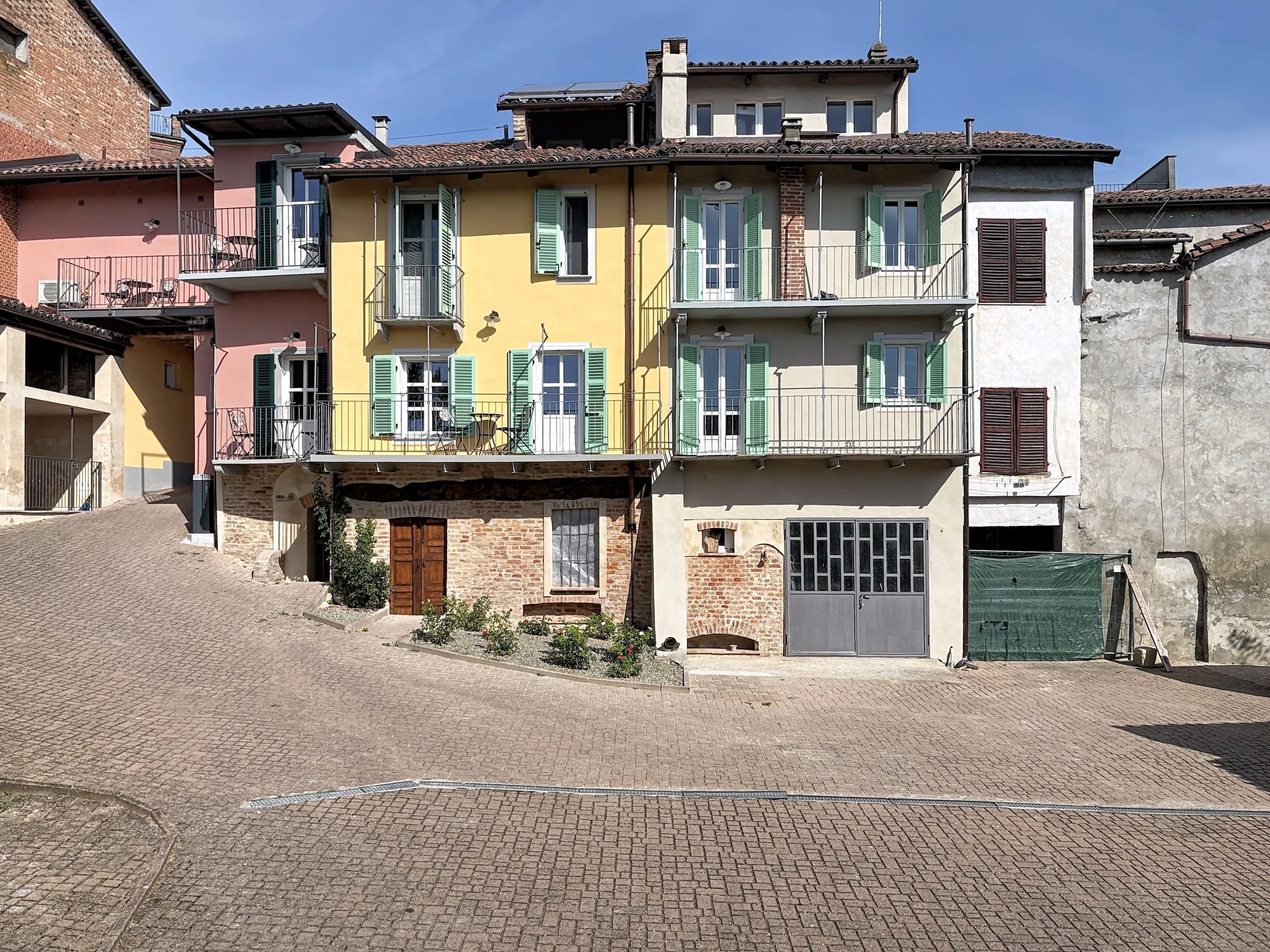 Une rangée de bâtiments aux toits de tuiles de terre cuite, dotés de balcons, une rue pavée et un petit espace jardin.