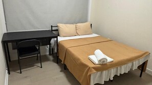 Ein kleines Zimmer mit einem Einzelbett, einem Schreibtisch und einem Stuhl, einer beigen Tagesdecke und einem aufgerollten weißen Handtuch auf dem Bett.
