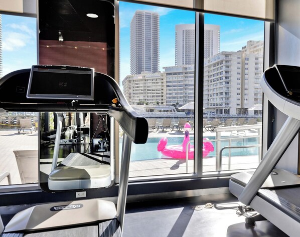 Academia com equipamentos de ginástica e vista para uma piscina com boia em formato de flamingo rosa.