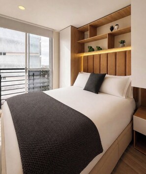 Una habitación de hotel moderna con una cama grande, un banco integrado al pie, una mesita de noche y un ventanal con vistas a la ciudad.