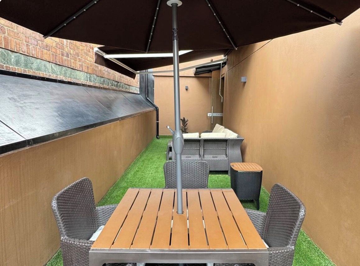 Un patio exterior con mesa de madera y sillas de mimbre, una parrilla de barbacoa y una zona de estar cubierta.