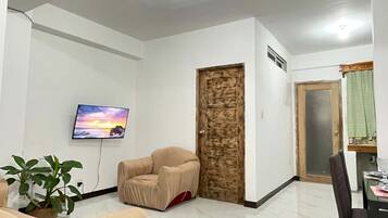 A living room with a flat-screen TV, a covered sofa, a dining table with chairs, a wooden door, and a potted plant.