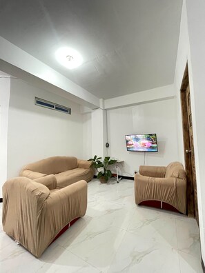 A room with a beige sofa, a television on the wall, and a potted plant.