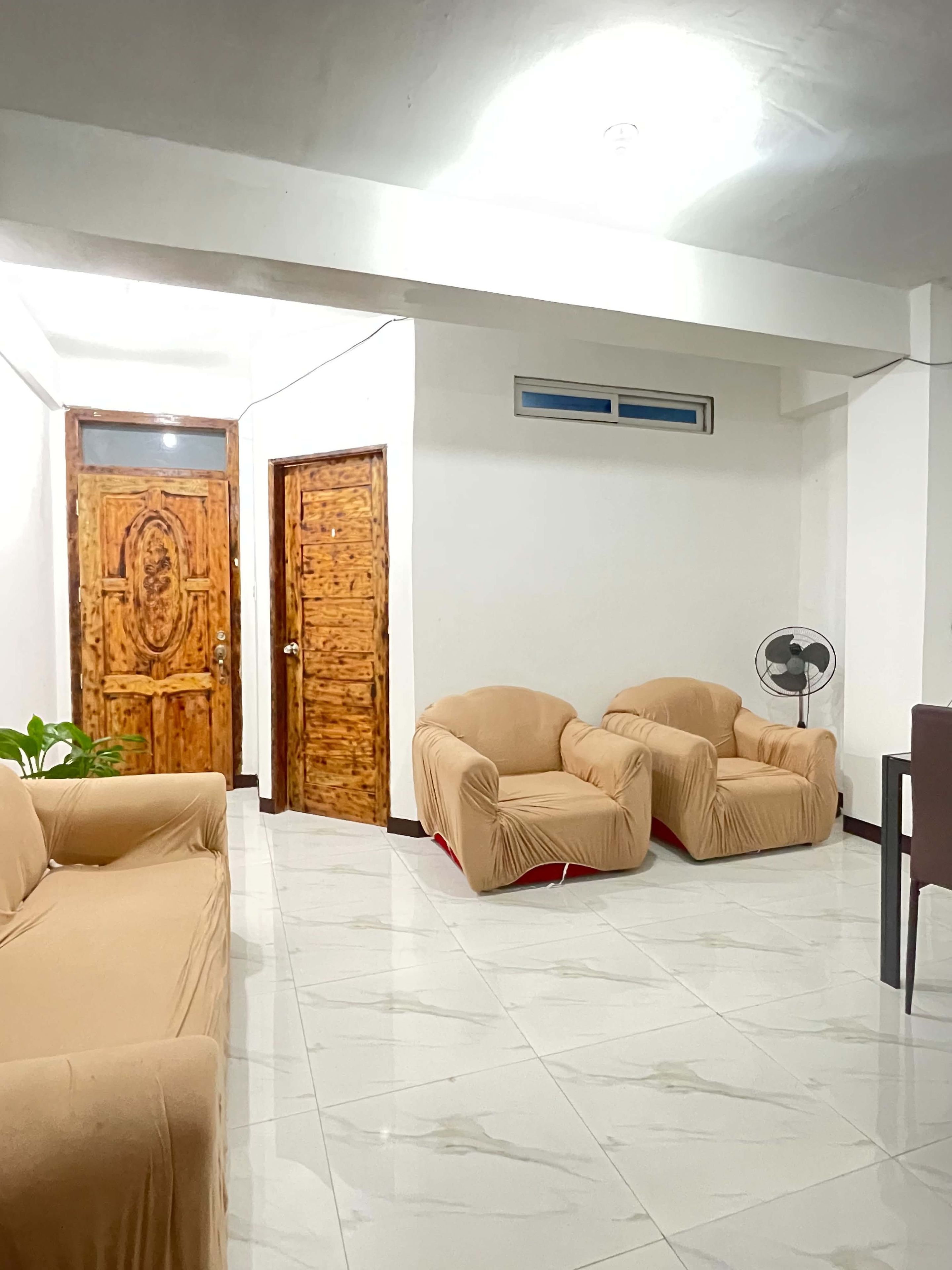 Una habitación con un sofá color crema, dos sillones color crema, una puerta de madera con panel de vidrio, una pared blanca con ventana y un ventilador de techo.