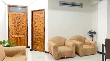 Una habitación con un sofá color crema, dos sillones color crema, una puerta de madera con panel de vidrio, una pared blanca con ventana y un ventilador de techo.