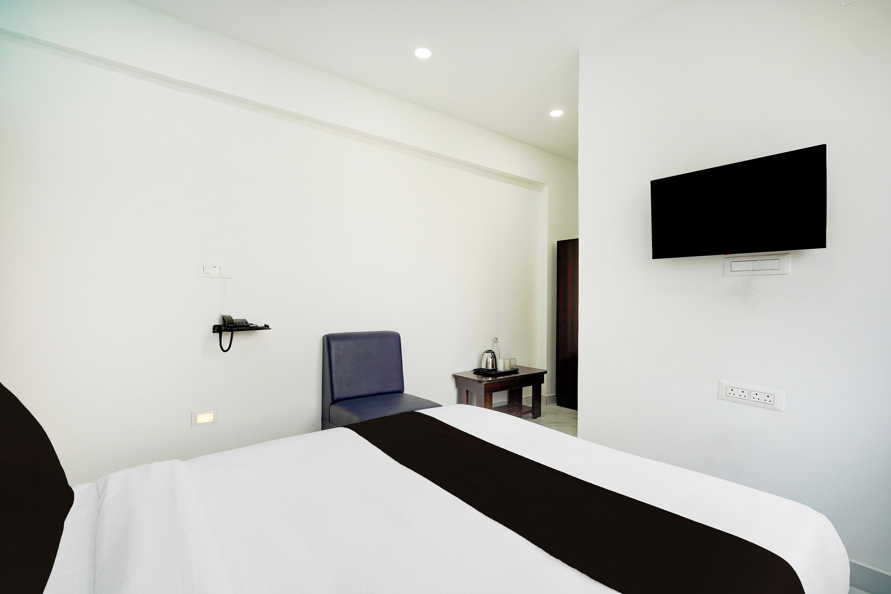 Habitación de hotel con cama, una silla, una mesita con una botella y vasos, teléfono, televisor montado en la pared y toma de corriente.