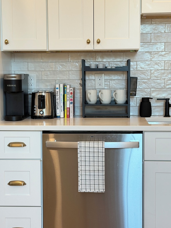 A kitchen with white cabinets, a coffee maker, a toaster, a dish rack with mugs, and a dishwasher.