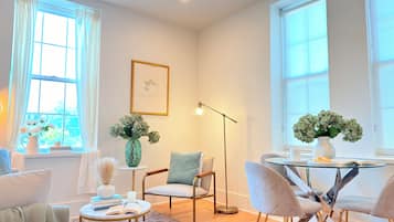 A modern living room with a sofa, chairs, a coffee table, and a dining table with chairs. There are windows with curtains, a floor lamp, and a vase with flowers.