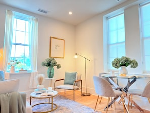 A modern living room with a sofa, chairs, a coffee table, and a dining table with chairs. There are windows with curtains, a floor lamp, and a vase with flowers.