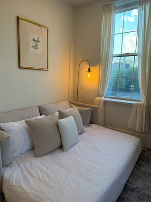 Une chambre à coucher avec un lit, un tableau encadré au mur, une lampe sur pied et une fenêtre avec des rideaux.