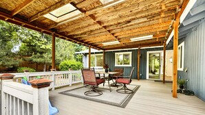 Una veranda coperta con un pavimento in legno, un tavolo con sedie e vista sul verde circostante.