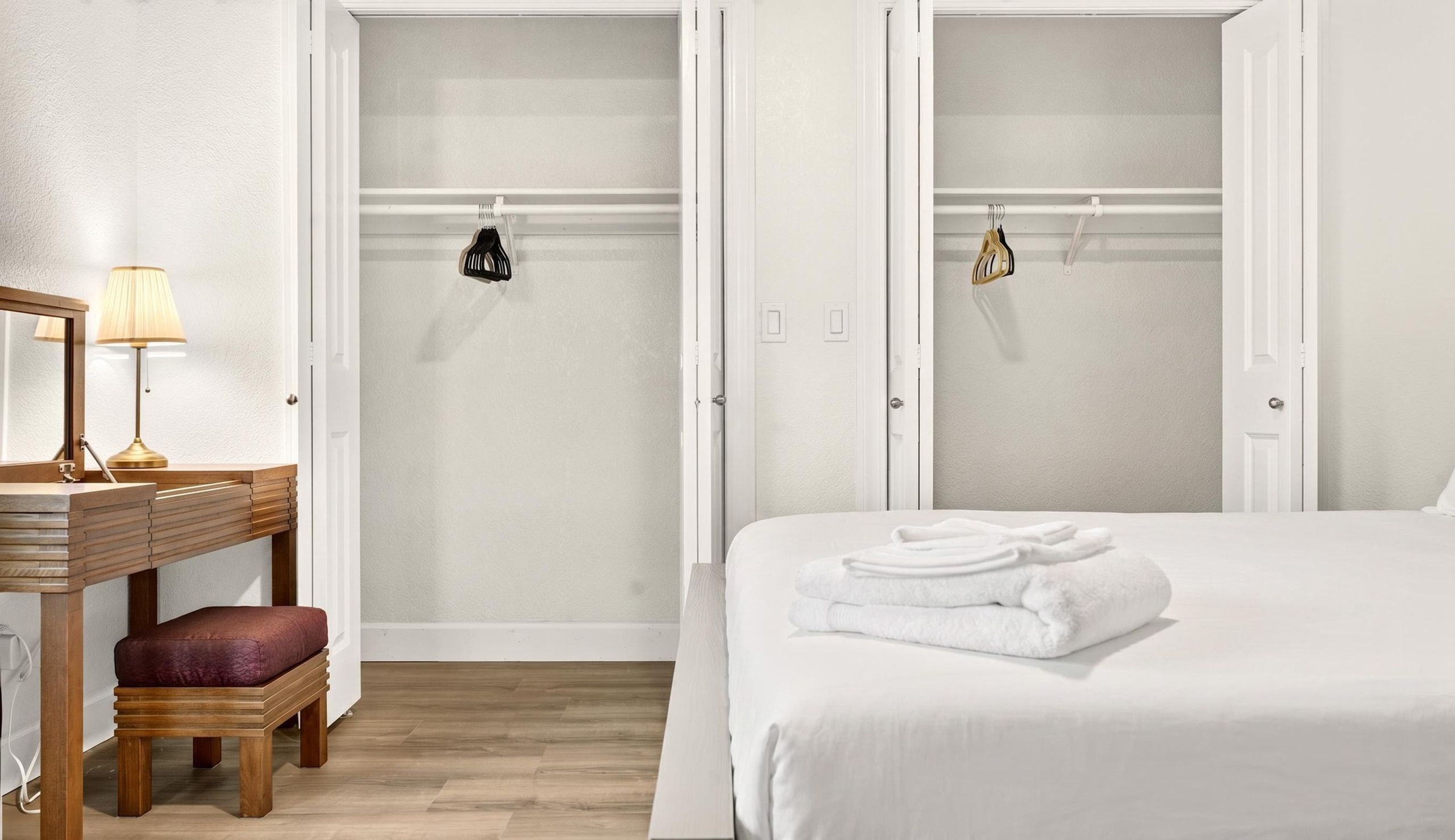 A hotel room with a bed, a bedside table with a lamp, a wooden bench, and a closet with hanging clothes.