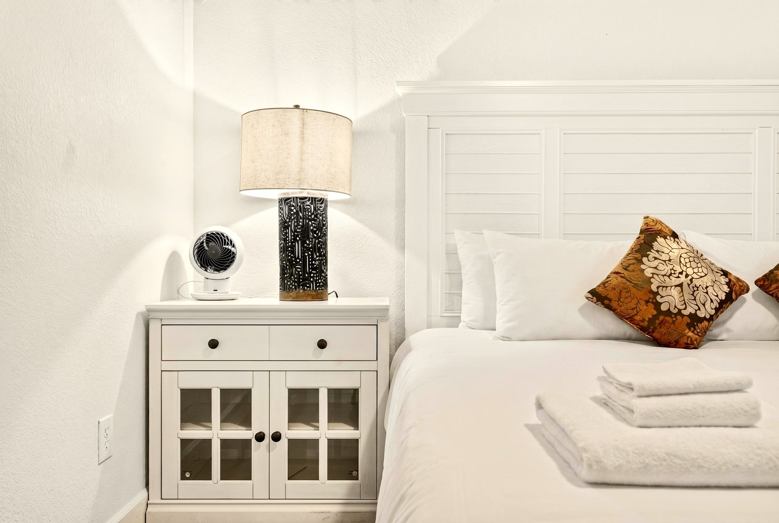 A neatly arranged bedroom with a white bed, a nightstand with a lamp, and a decorative item.