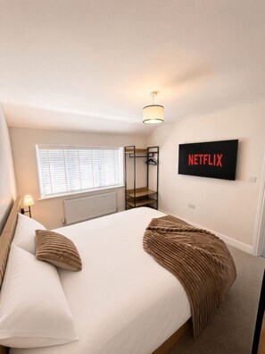 Ein Schlafzimmer mit Bett, einer Hängeleuchte, einem Fenster mit Jalousien, einem Heizkörper, einem Kleiderständer und einem an der Wand montierten Fernseher mit Netflix-Logo.