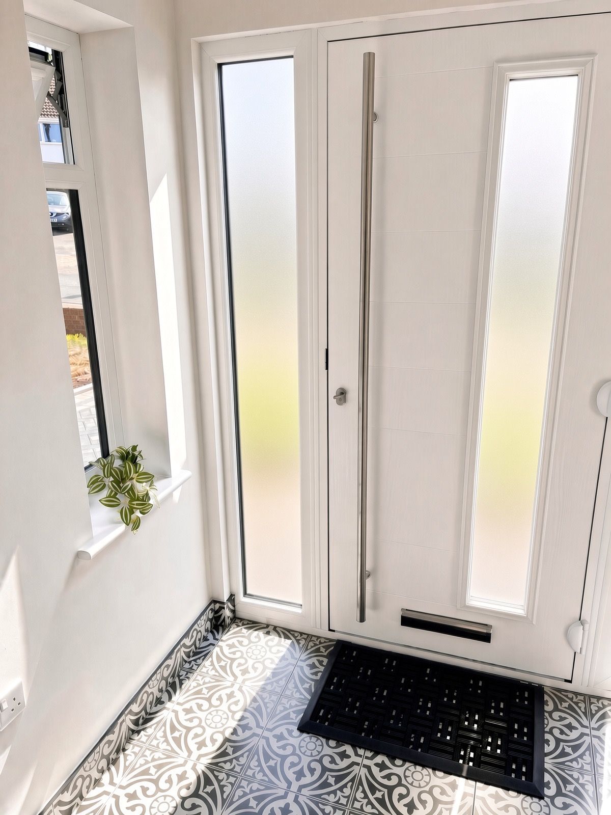 Ein Zimmer mit einer weißen Tür, einem Fenster mit Pflanze, gemusterten Bodenfliesen und einer schwarzen Matte am Eingang.
