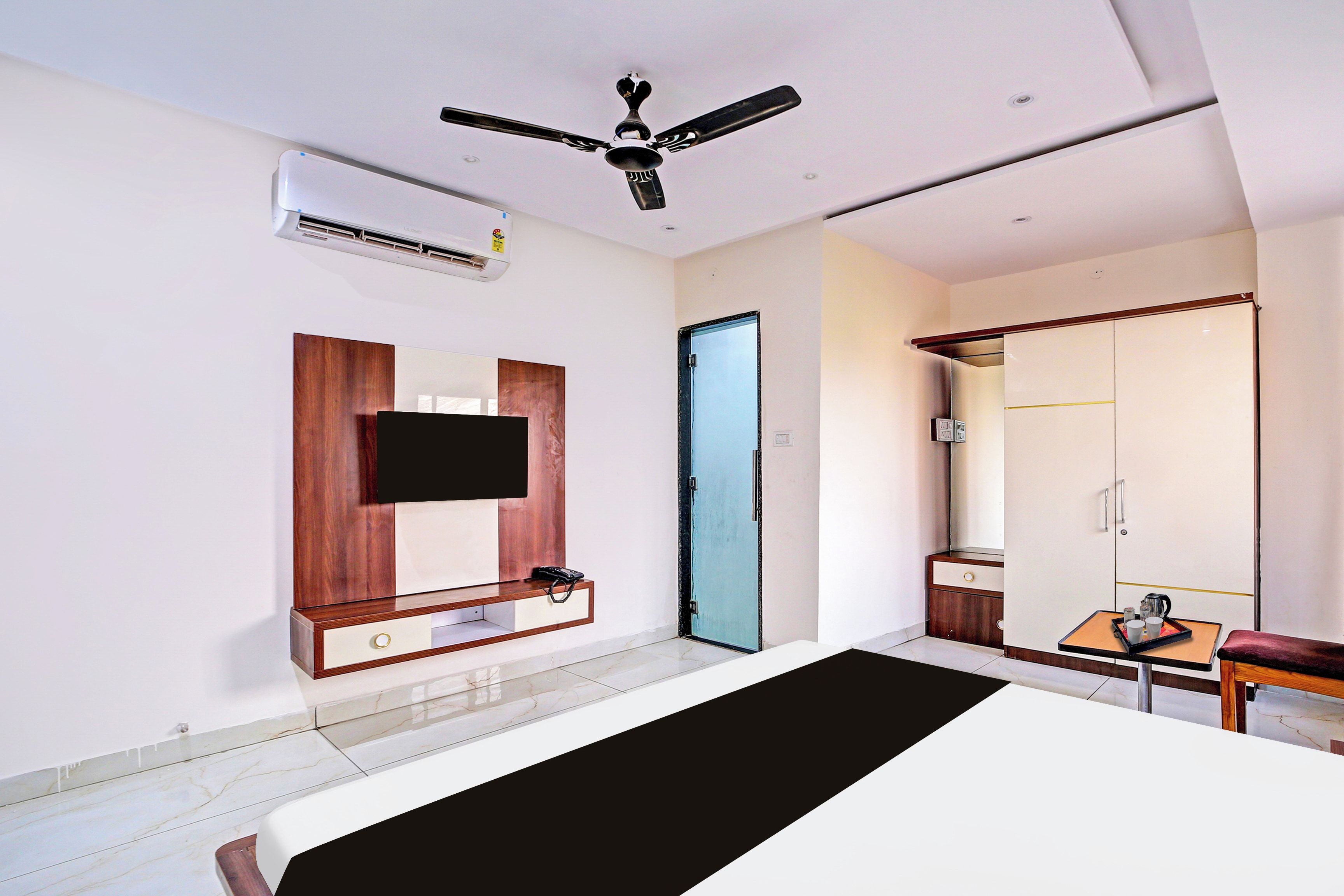Habitación de hotel con cama, televisor de pantalla plana montado en un panel de madera, ventilador de techo, equipo de aire acondicionado, armario y una pequeña mesa con bandeja.