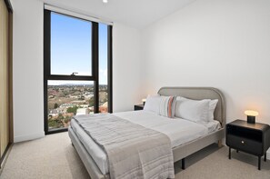 A modern bedroom with a large bed, a nightstand, a bedside lamp, and a view of a cityscape through a large window. - Balcony 3-bed Apartment Near Albert Park (Melbourne)