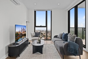 A modern living room with a large TV, a sofa, a coffee table, and a view of a cityscape through large windows. - Balcony 3-bed Apartment Near Albert Park (Melbourne)