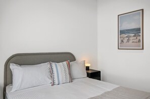 Ein Bett mit grauem Kopfteil, weißer Bettwäsche und gestreiften Kissen. Ein Nachttisch mit Lampe steht neben dem Bett. An der Wand hängt ein gerahmtes Bild von einer Strandlandschaft.