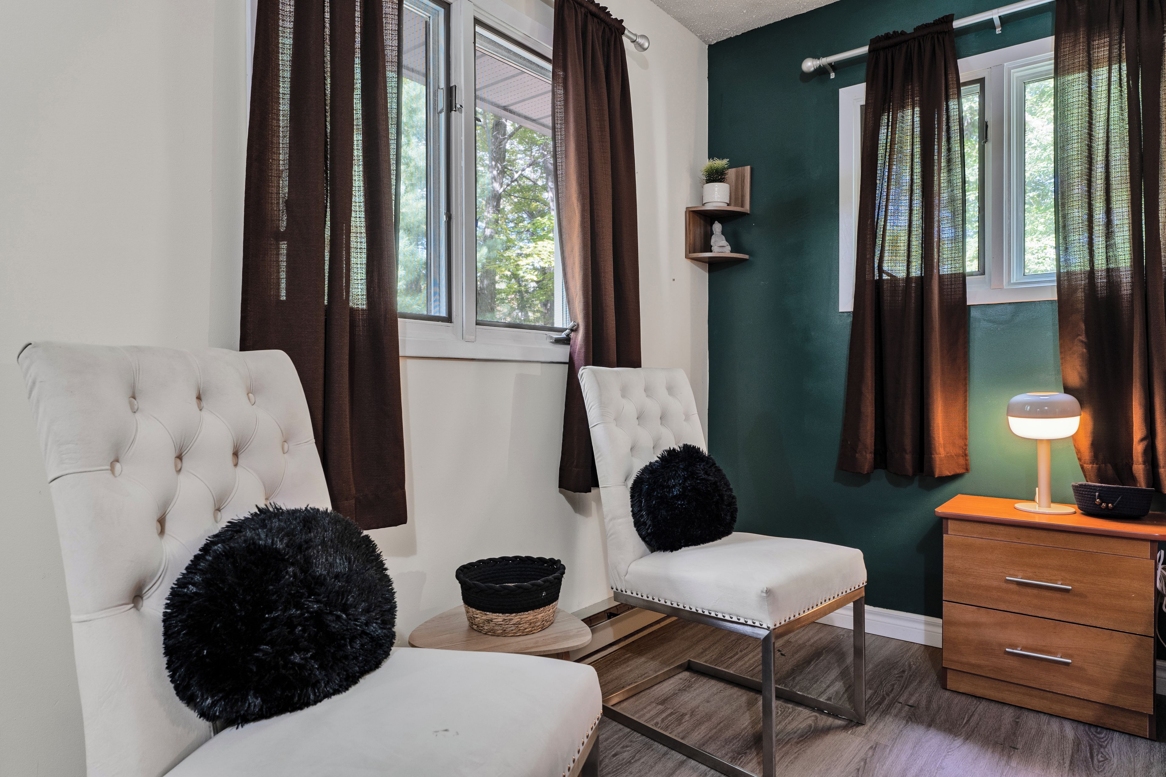Ein Zimmer mit zwei weißen, gepolsterten Sesseln, einem Holznachttisch mit Lampe und einem kleinen Regal mit Dekorationsobjekten.