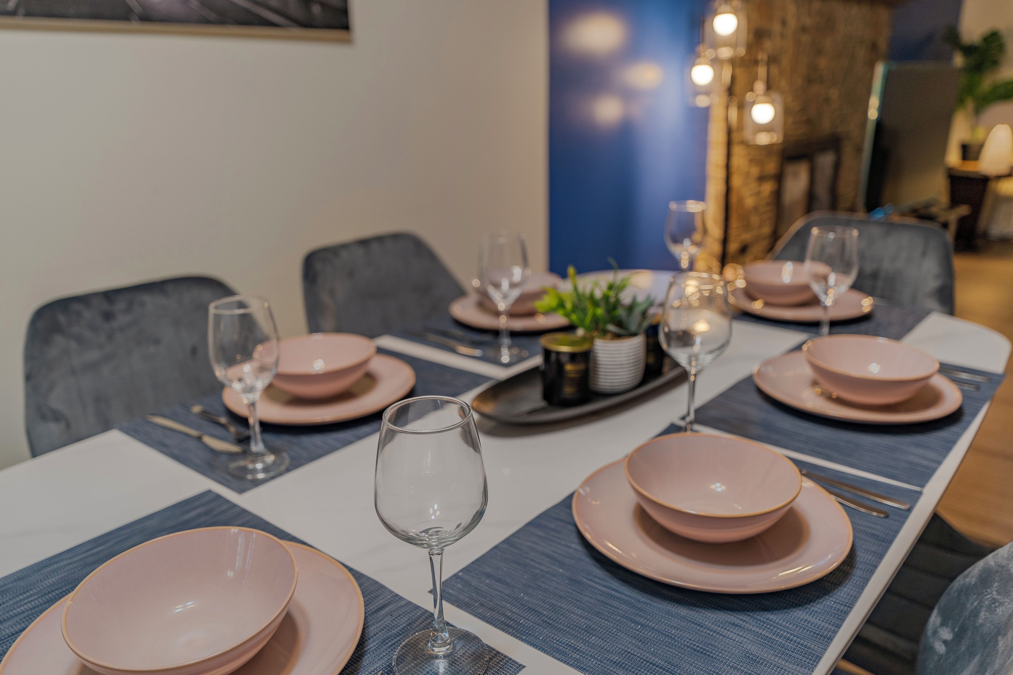 Una mesa de comedor dispuesta con platos rosas, vasos y cubiertos, con un mantel individual azul y un pequeño centro de mesa con planta.
