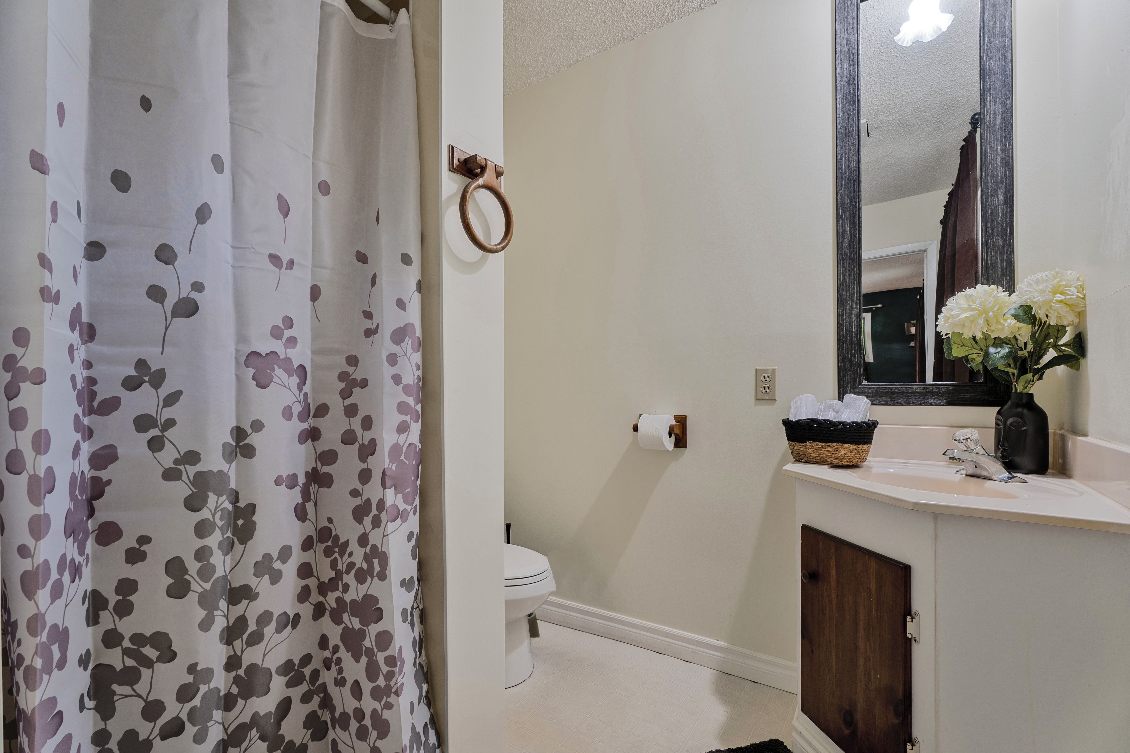 A bathroom with a toilet, a sink with a mirror, a shower curtain, and a vase with flowers on the sink.