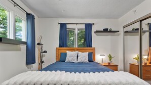 Un dormitori amb un llit, una tauleta de nit, una làmpada, una estant amb objectes decoratius i una finestra amb cortines.