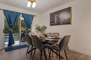 En spiseplads med et bord dækket til fire personer, et stort vindue med gardiner og et billede i ramme på væggen.