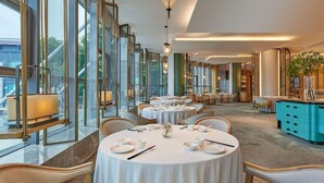 Un comedor de hotel con mesas redondas dispuestas para el servicio, ventanas amplias y vistas al exterior.
