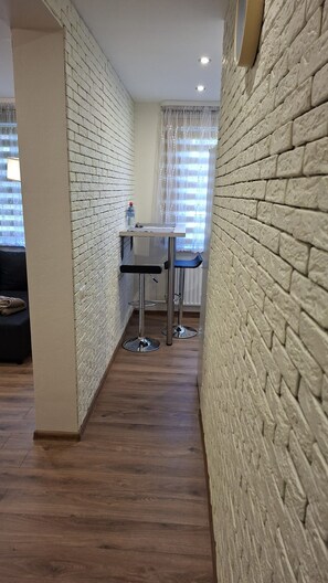 Un couloir étroit, avec un mur de briques d’un côté et une fenêtre avec des rideaux de l’autre. Au centre, on trouve une petite table et une chaise.