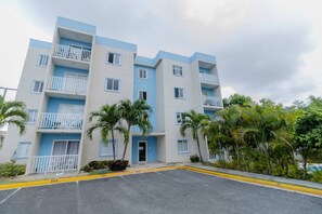 Un edificio residencial de varios pisos con balcones, zona de estacionamiento y vegetación circundante.