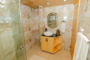 Un baño con cabina de ducha de vidrio, tocador de madera con lavabo blanco y pared con azulejos de mármol.