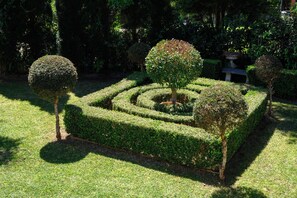 Um jardim formal com um labirinto de sebes geométricas, árvores bem aparadas e um banco de pedra.