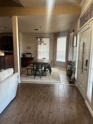 A spacious dining area with a wooden table and chairs, a kitchen in the background, and a large window with blinds. - Soothing 3-bedroom house with AC in charming Lawton (Lawton)
