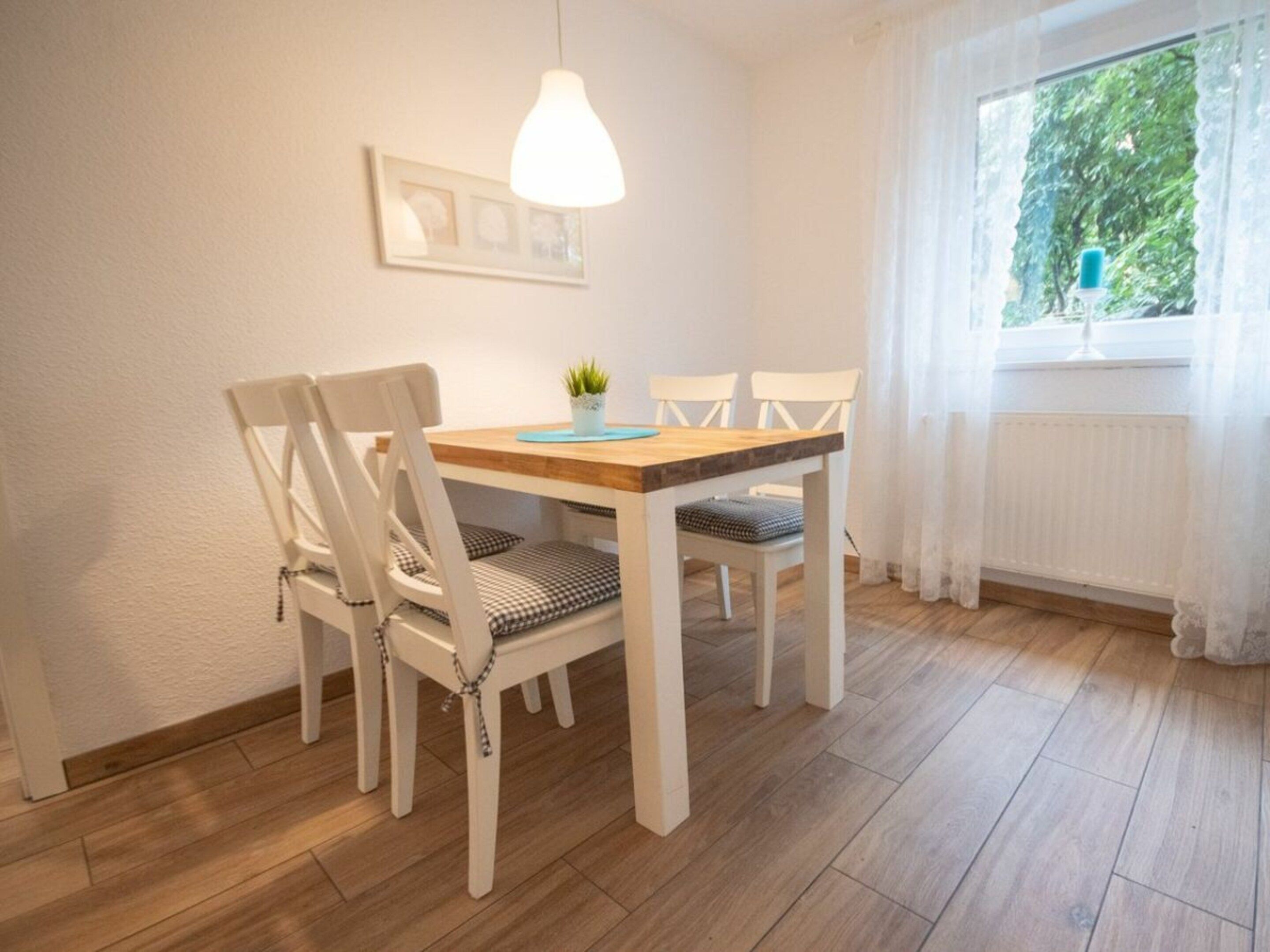 Un espace repas avec une table en bois et des chaises blanches, un luminaire suspendu et une fenêtre donnant sur un paysage verdoyant.