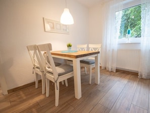 Ein Essbereich mit einem Holztisch und weißen Stühlen, einer Hängelampe sowie einem Fenster mit Blick ins Grüne.