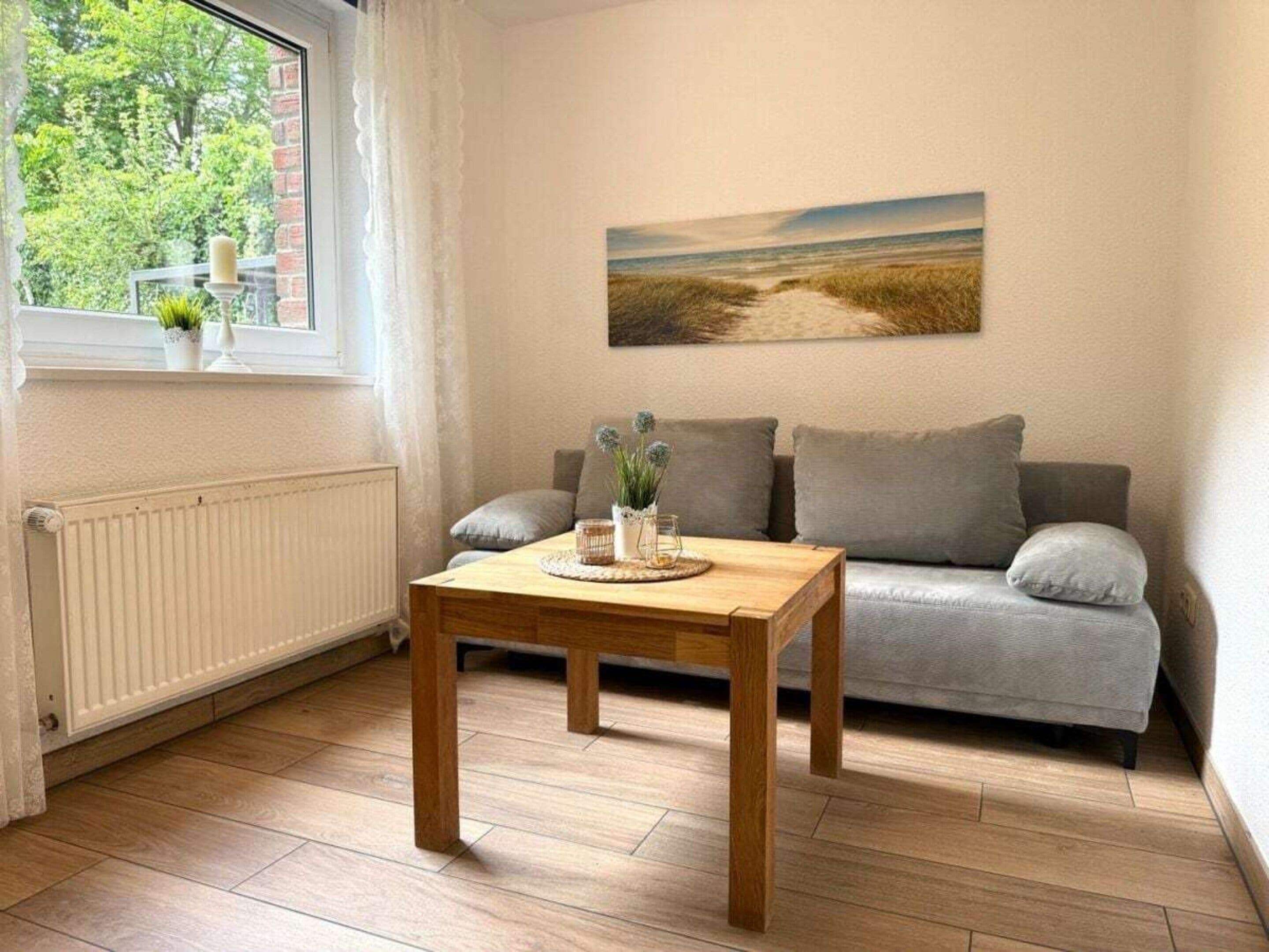 Ein gemütliches Wohnzimmer mit Holztisch, grauem Sofa und einem Fenster mit Blick auf Grünflächen.