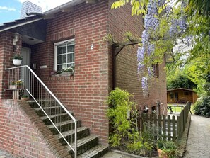 Una casa de ladrillo con una escalera, una valla de madera y un jardín con flores moradas.