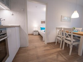 Una cocina moderna con horno empotrado, zona de comedor con mesa de madera y sillas blancas, y vista a una playa a través de una puerta abierta.