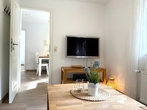 Ein Zimmer mit einem an der Wand befestigten Fernseher, einem Holztisch mit geflochtener Unterlage und einer Topfpflanze.