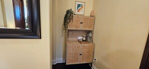 A hallway with a framed mirror, a rattan cabinet with kitchen items, and a plant.