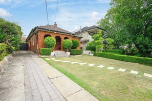 A residential property with a paved driveway, a brick building with arched windows, and well-maintained lawns and hedges. - Linley Point Luxury 5BR Home with Pool BBQ (Linley Point)