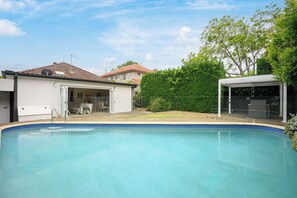 Una piscina con zona cubierta, una casa con garaje abierto y un seto al fondo.