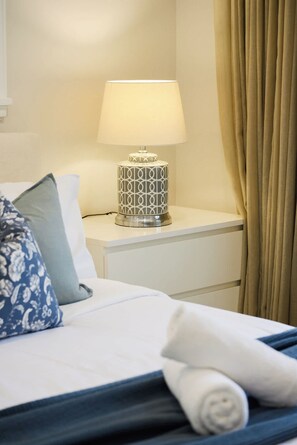 Un lit bien fait, recouvert d’une couvre-lit blanche et bleue, agrémenté d’un coussin décoratif et d’une lampe de chevet au motif géométrique, posée sur une table de chevet blanche.