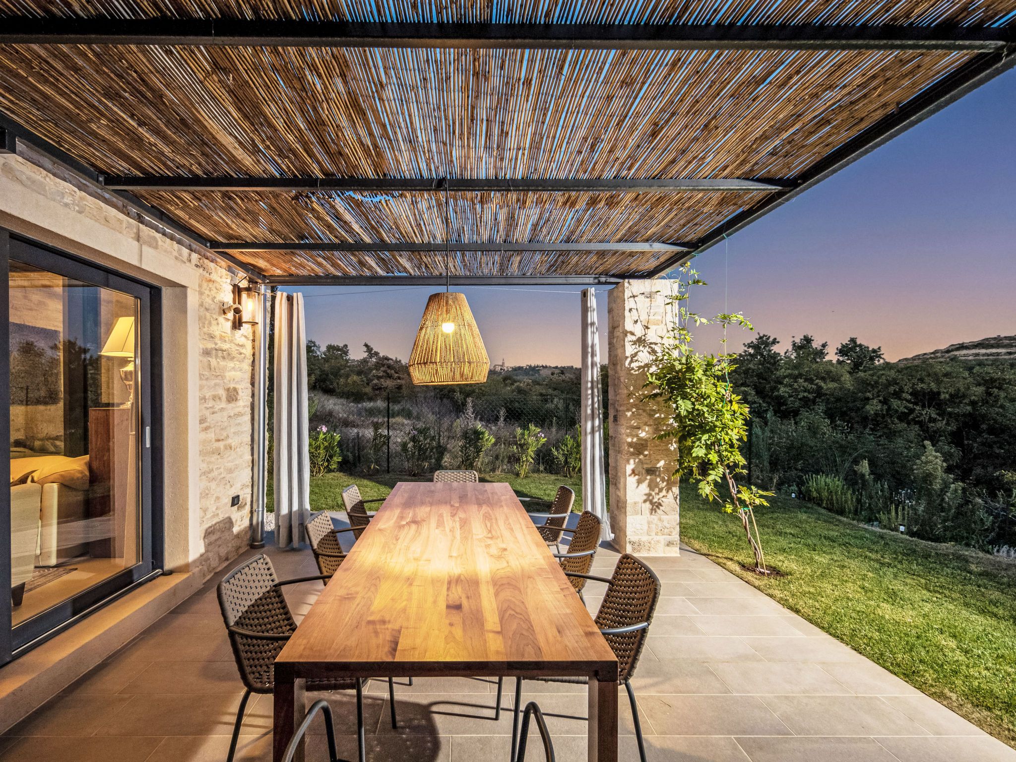 An outdoor patio with a wooden table and chairs, a hanging light fixture, and a view of the surrounding landscape.