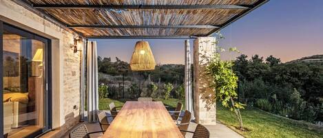 An outdoor patio with a wooden table and chairs, a hanging light fixture, and a view of the surrounding landscape.