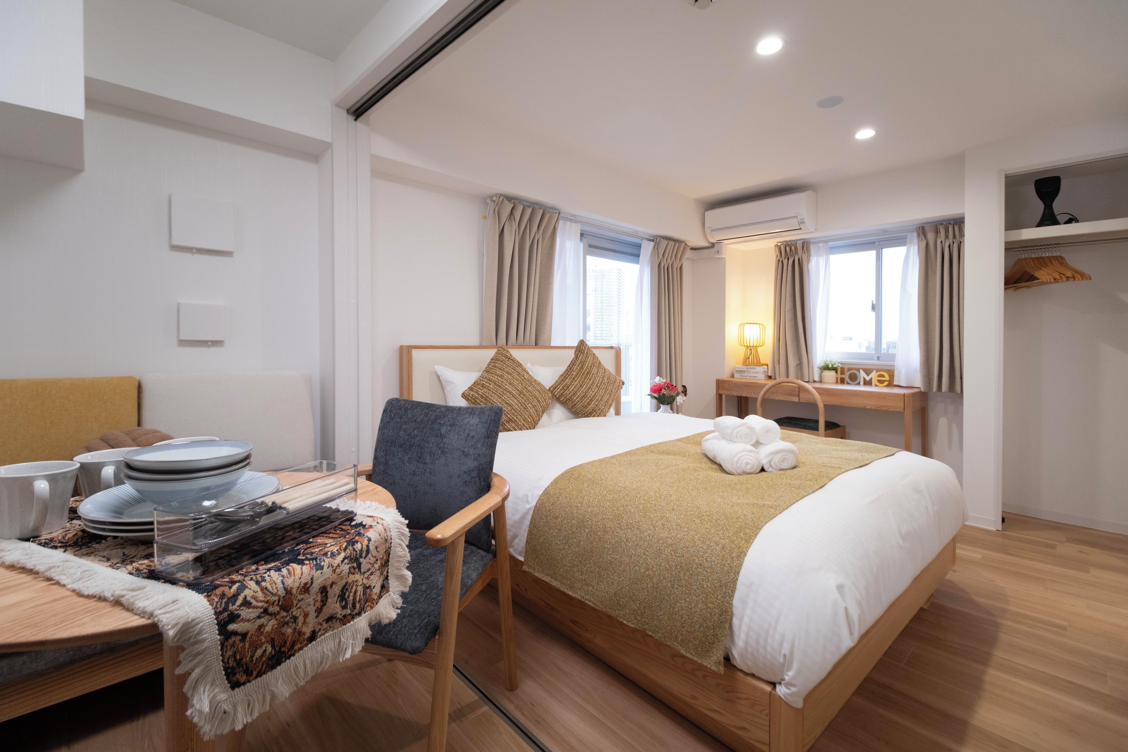 A hotel room with a bed, a desk with a chair, a small table, and a window with curtains.