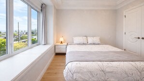 Un dormitorio con un ventanal grande, una cama con una colcha estampada de árboles, una lámpara de noche y una puerta blanca.