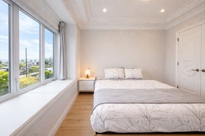 Une chambre avec une grande fenêtre, un lit recouvert d’une couvre-lit à motifs d’arbres, une lampe de chevet et une porte blanche.