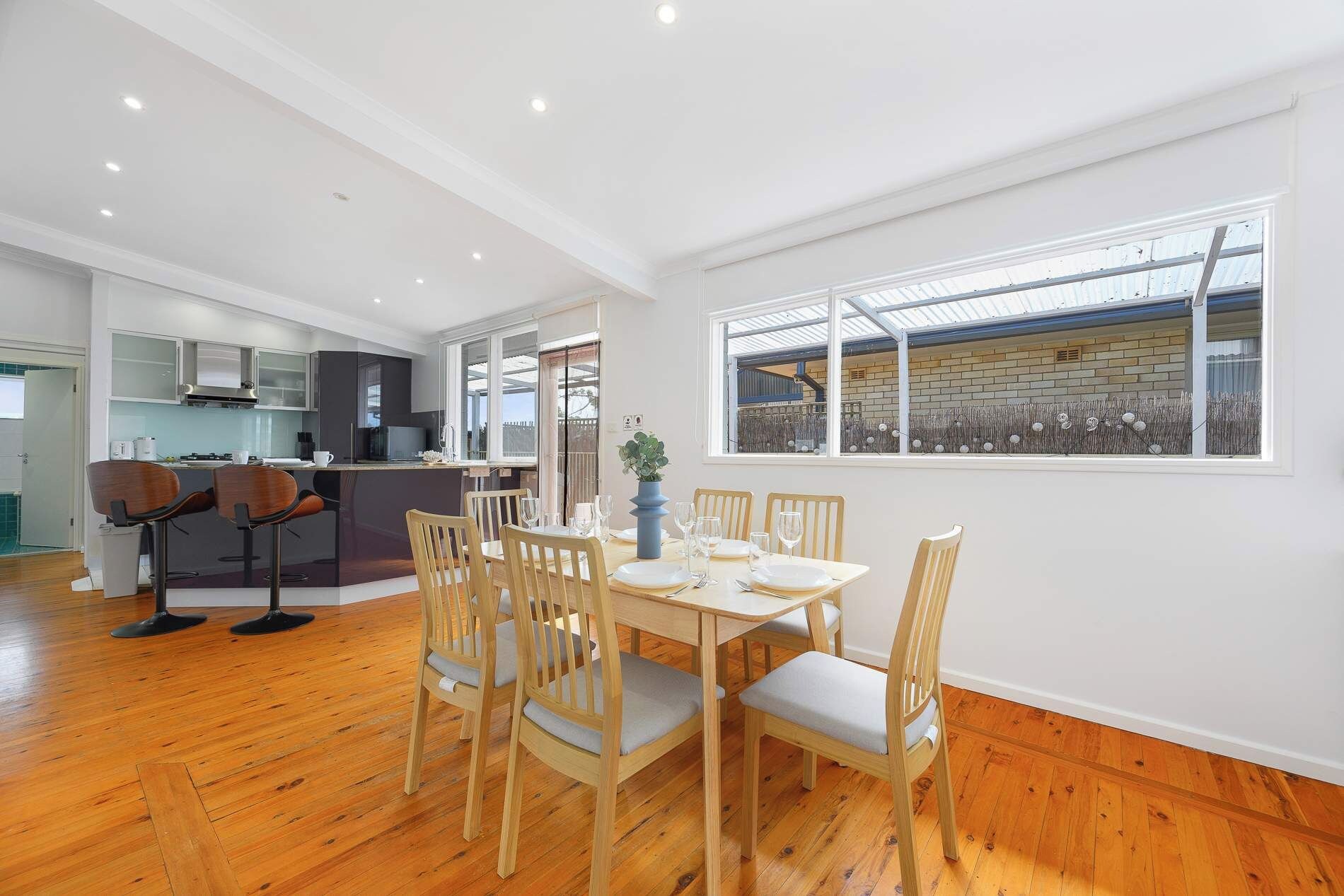 A dining area with a wooden table and chairs, a kitchen in the background, and a large window with a view.